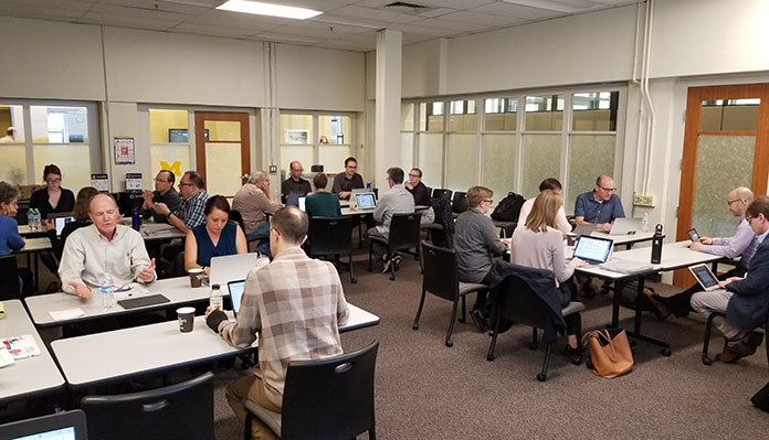 A room with four groups of three to six people seated around tables, most with laptops in front of them.