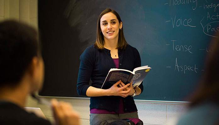 A young female professor smiles and looks out toward her class while holding a textbook and standing in front of a chalkboard with the words "Voice, Tense, and Aspect" visible. The back of a male student's head can be seen in the left foreground.