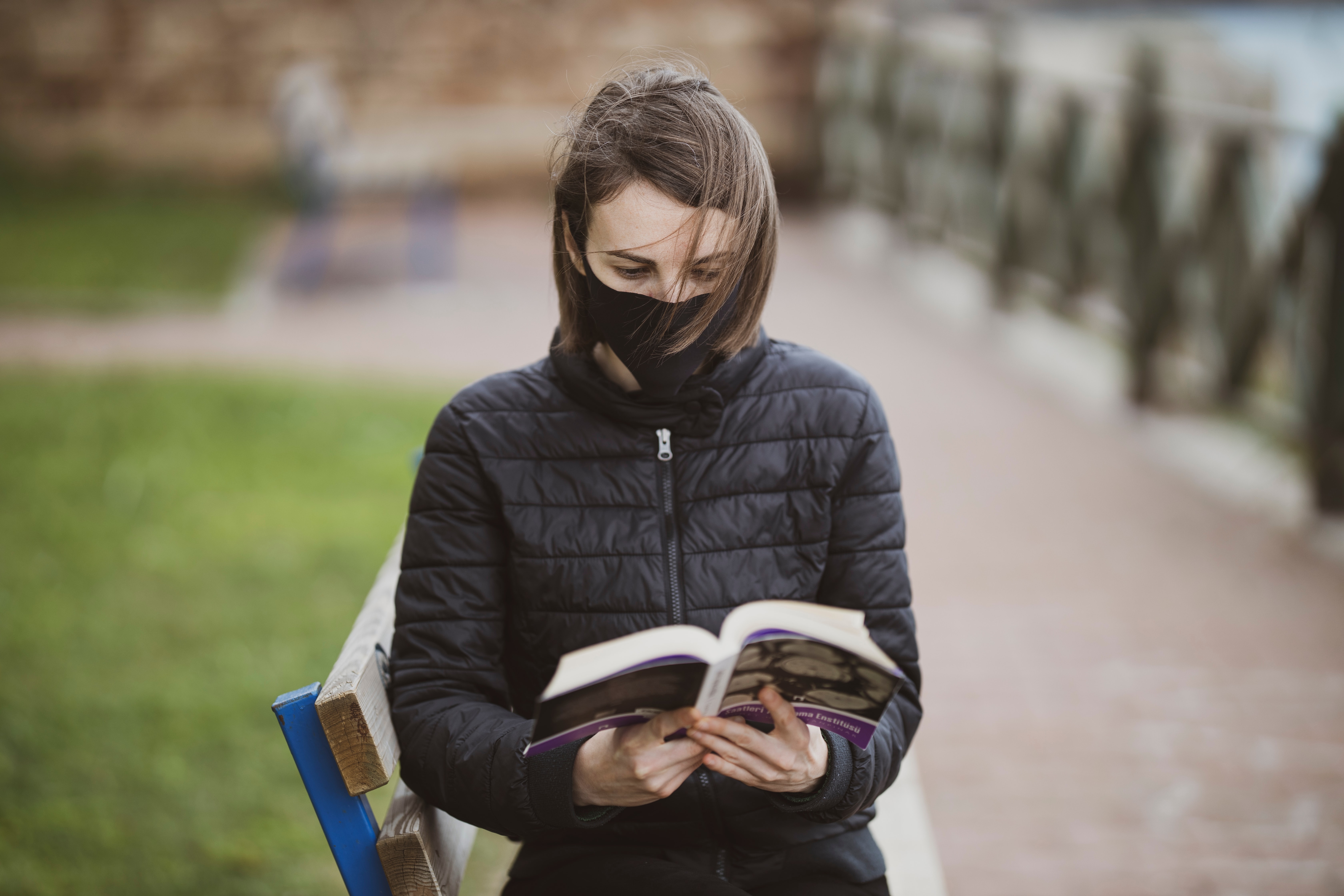 Picture of white woman in black coat and black face mask sitting on isolated park bench reading a book