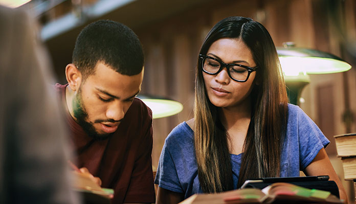 A photograph of two students of color studying from a book together in what appears to be a library or study hall.