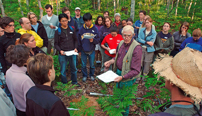 A male instructor holds a sheet of paper while standing in a forest surrounded by college students.